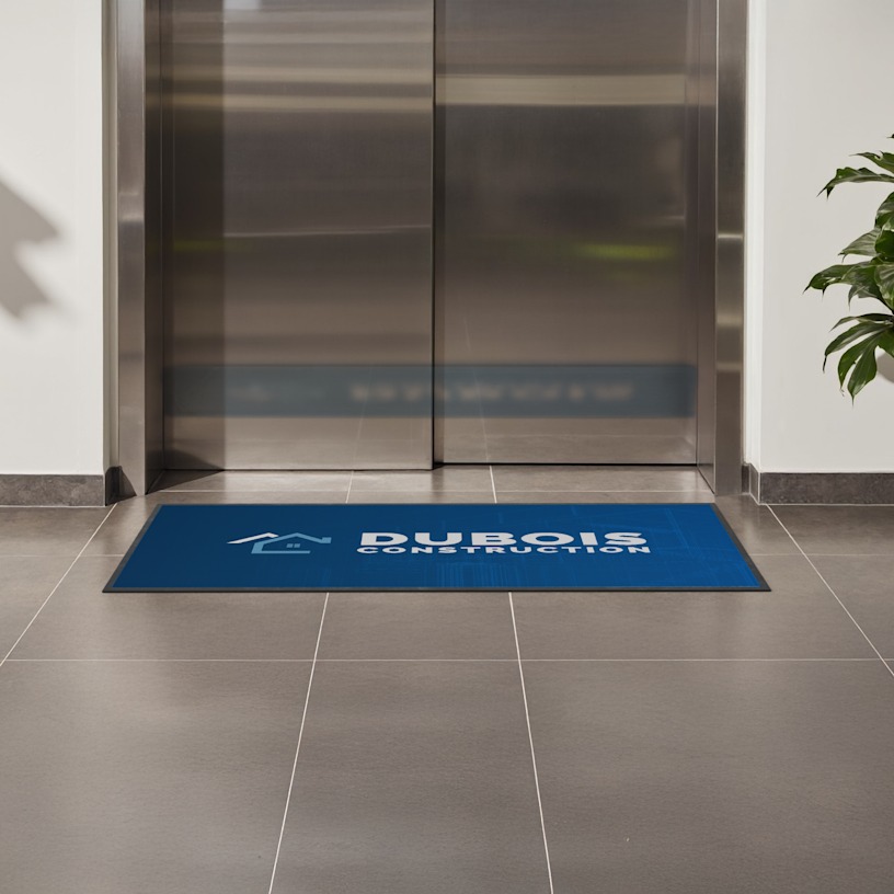 Tapis de sol arborant le logo d’une entreprise de construction, placé devant un ascenseur.