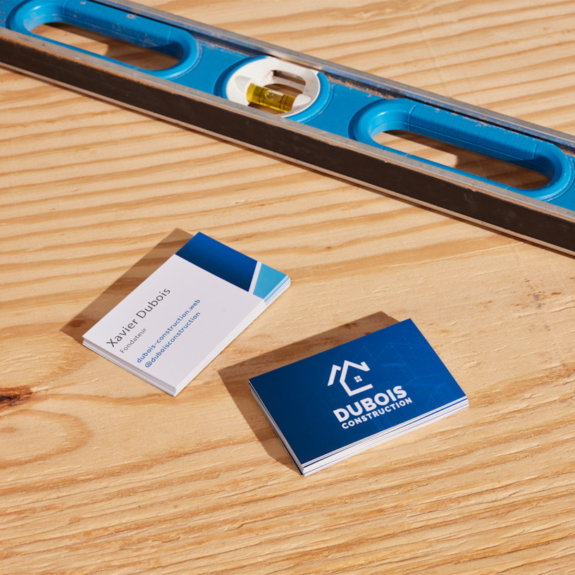 Piles de cartes de visite express 24h pour une entreprise de construction posées sur un comptoir en bois, à côté d’un niveau à bulle.