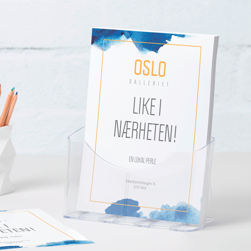 En A4 brosjyreholder i plast med flyere for et galleri på et skrivebord ved siden av en penneholder.