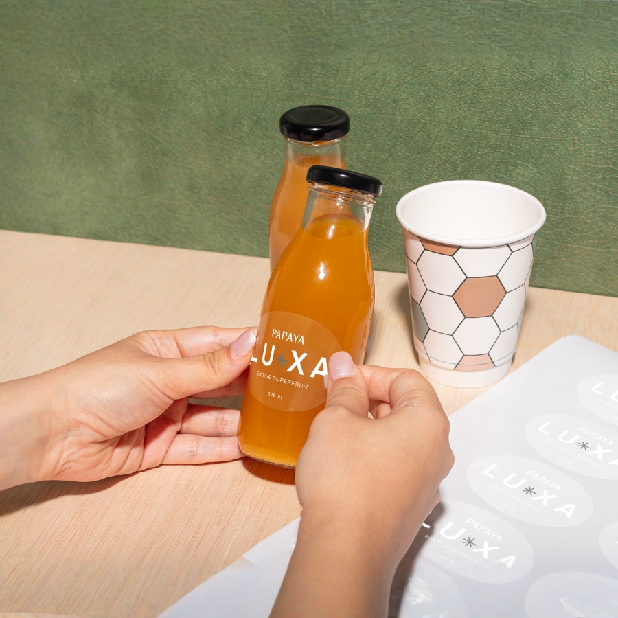 Een hand plaatst een ronde, bedrukte Luxa-sticker op een fles op een houten tafel, naast een stickervel.