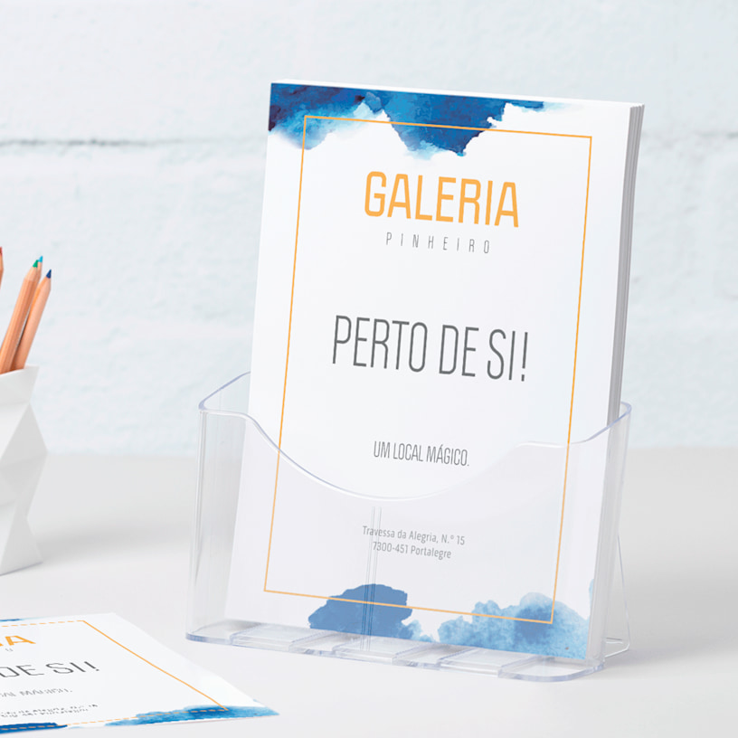 Um suporte de folhetos em plástico de tamanho A4 com flyers de uma galeria pousado numa secretária junto a um porta-lápis.