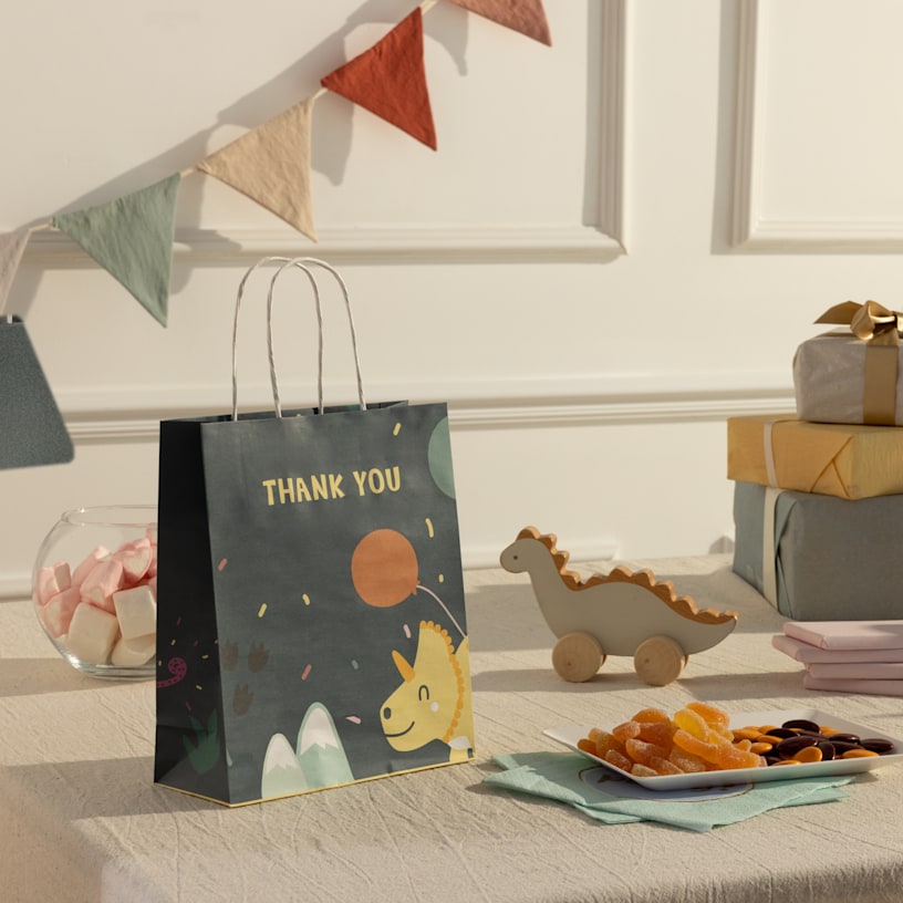 Uma mesa de aniversário decorada com um saco para presentes personalizado, com tema de dinossauros e etiqueta com a palavra "Obrigado".
