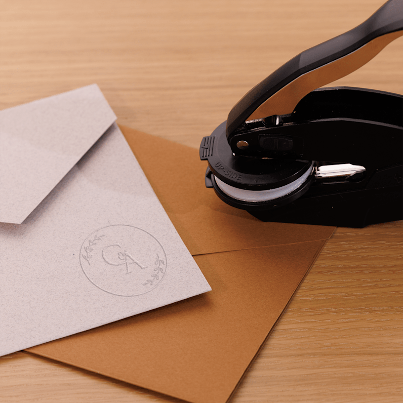 Um carimbo de alto relevo ao lado de envelopes em papel kraft com um selo branco com monograma, dispostos sobre uma superfície de madeira.