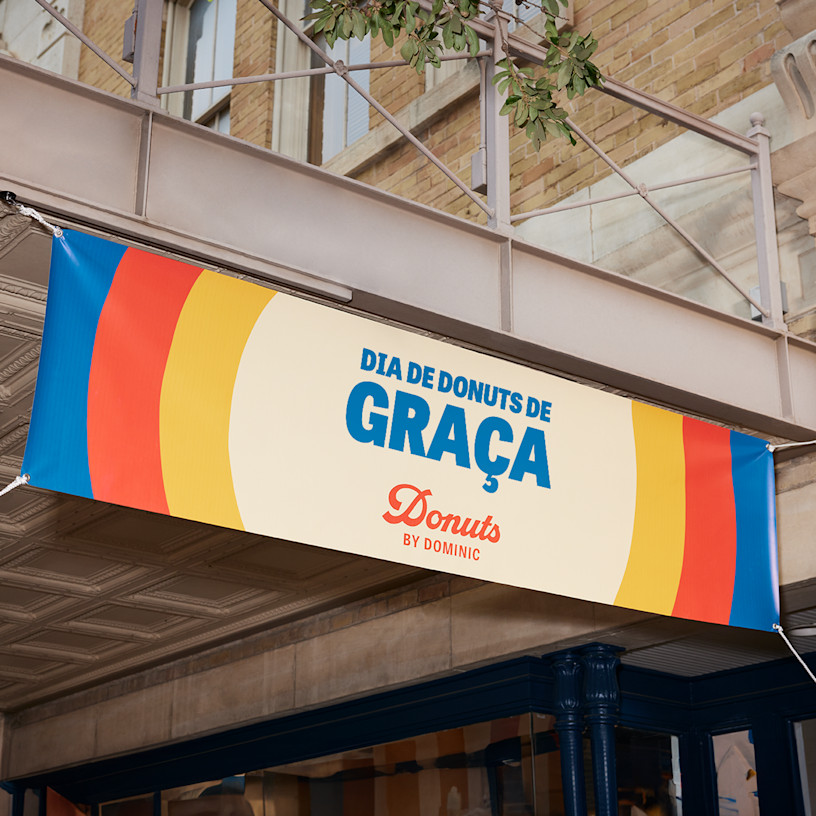 Lona publicitária com a mensagem “FREE DONUT DAY!” escrita com letras grandes e a negrito, pendurada no exterior de uma loja de donuts.