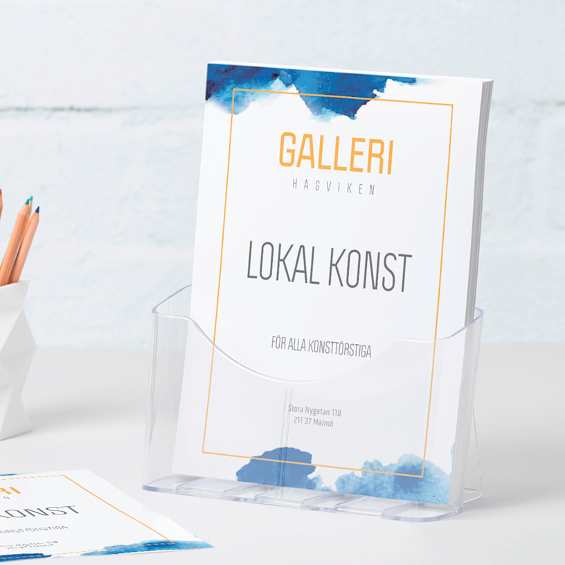 Ett broschyrställ i plast i A4-storlek med flyers för ett galleri står på ett bord bredvid en pennhållare.