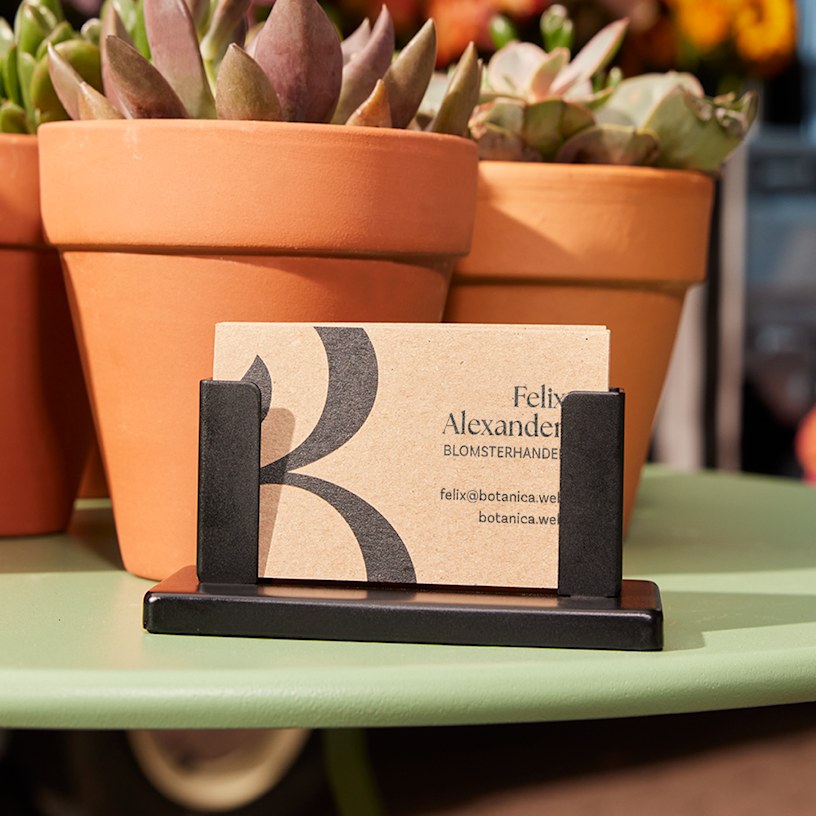 En hög visitkort av kraftpapper för en blomsterbutik visas i en svart korthållare, bredvid suckulenter.