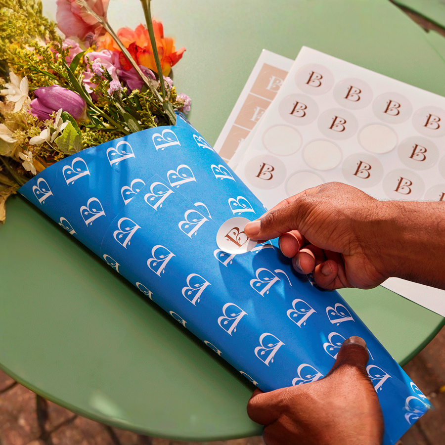 A circle sticker promoting a flower shop, being applied onto matching bouquet packaging.