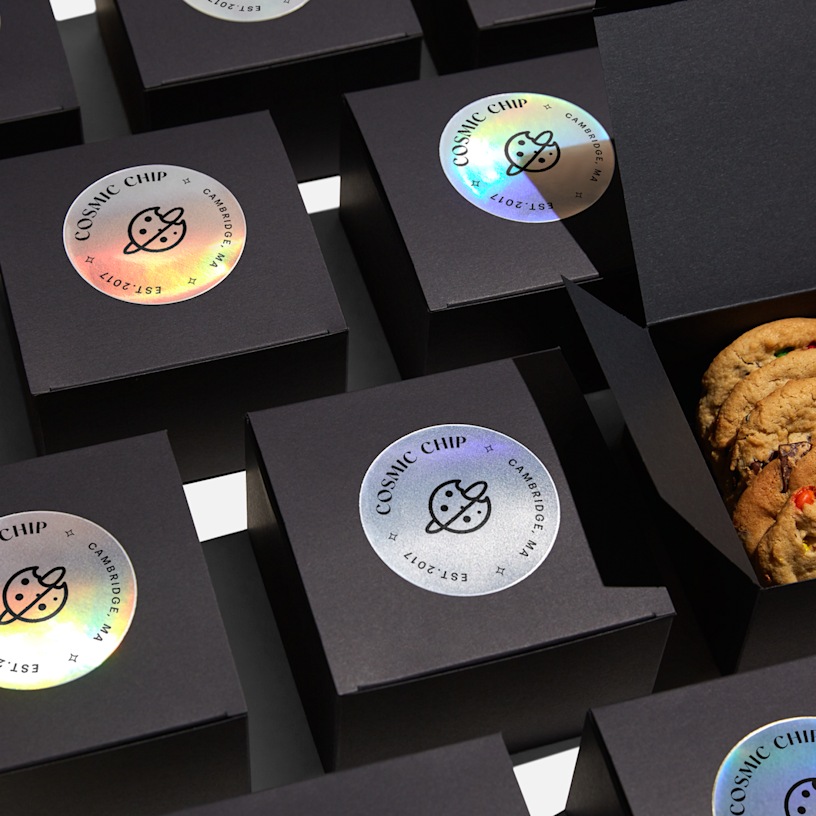 Black boxes neatly lined up in rows with circle holographic paper sticker labels applied to the top of each. One box is opened revealing cookies inside