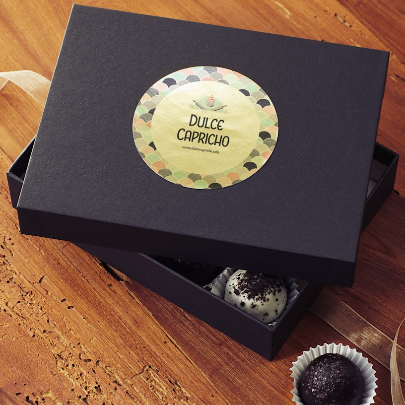 A black box sitting on a wooden table with the cover off revealing bon bons inside. The lid has a circular gold paper label applied to it.