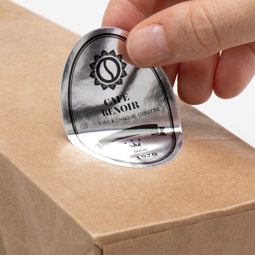 A bag of coffee on a gray background with a hand applying a silver paper oval label to the front.