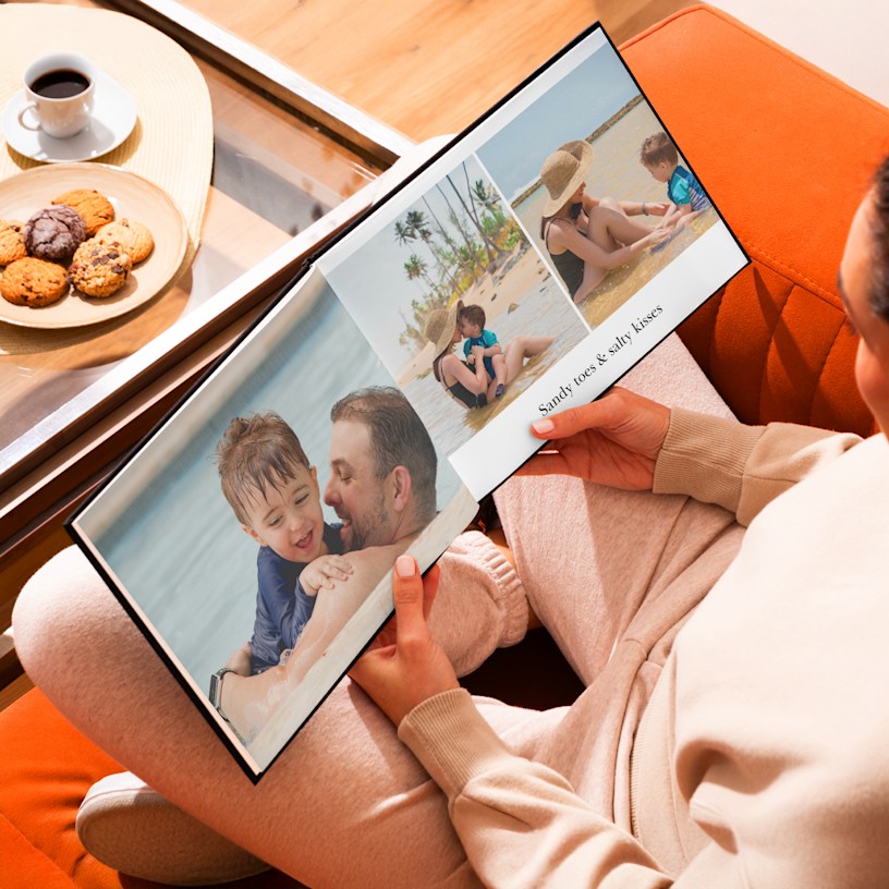 A pair of hands holding an open photo book over a coffee table. The photos show a family at the beach.
