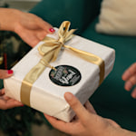 A person handing a wrapped present with a Christmas ornament looped around the ribbon. The ornament features a stylised Christmas greeting.