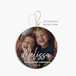 A Christmas ornament featuring a photo of two children, along with a festive greeting. An arrow points to the pre-drilled hole and gold string.