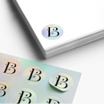 A sheet of custom stickers next to a stack of white papers. A holographic round sticker with the letter B and a leaf design is on the papers.