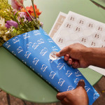 A person is sealing a bouquet of blue flowers with a custom-made circle sticker featuring a ‘B’ logo on a green table adorned with flowers.