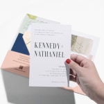 A hand holding a wedding invitation printed with a bold font and a simple black and white color palette. 