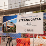 En banderoll från ett byggbolag fäst på stängslet vid en byggarbetsplats, med information om den nya fastigheten.