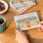A pair of hands resting on a table holding a painted edge holiday greeting from a floral business. 