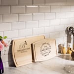 Two custom cutting boards leaning against a kitchen backsplash, one customized for a winery and the other with a family name. 