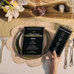Deux élégants menus noirs et dorés pour un 50e anniversaire, présentés sur une table dressée avec soin, l’un posé sur une assiette et l’autre tenu par un invité. 