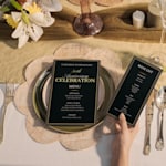 Two elegant black-and-gold 50th anniversary celebration menus displayed on a formally set dining table, with one menu on a plate and another held in a guest's hand. 