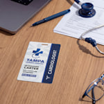 A cardiologist’s ID badge on a desk beside their laptop, glasses, pen and stethoscope.
