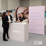 A fabric display wall promoting a cosmetics brand, placed behind a trade show counter with product displays.