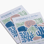 Deux cartes postales aimantées provenant d’un restaurant, arborant un graphisme coloré au fini brillant en relief.