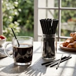 Un recipiente lleno de mezcladores de café plásticos color negro al lado de una taza de café con un mezclador de café negro adentro.