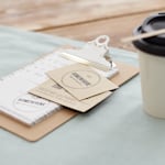 To visitkort i kraftpapir i standardstørrelse ligger ved siden af et papkrus med takeaway-kaffe.