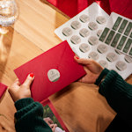 Eine Hand positioniert einen runden personalisierten Luxa-Aufkleber auf einer Flasche, die auf einem Holztisch steht; daneben ein Aufkleberbogen.