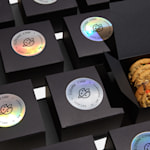 Black boxes neatly lined up in rows with circle holographic paper sticker labels applied to the top of each. One box is opened revealing cookies inside