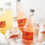 Various bottles of juice and yogurt cups arranged in a bucket of ice. Each bottle and yogurt cup is affixed with a clear plastic label.