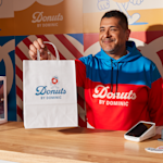 A smiling donut shop employee handing a branded kraft paper bag to a customer.