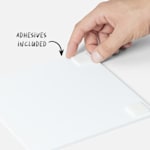 A hand placing a double-sided adhesive strip on the back of a photo tile with a note that adhesives are included with the tiles.