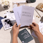 A pair of hands holding a wedding invitation printed with a bold font and a neutral colour palette.