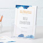 An A4 plastic leaflet holder with gallery flyers rests on a desk next to a pencil holder.