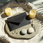 A black, custom envelope resting on a stone tray, surrounded by candles and pebbles.