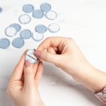 A pair of hands peeling off the backing of a blue, custom wax seal. 