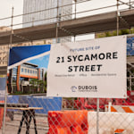 A banner for a construction company tied to a construction site fence, featuring details about the new building.