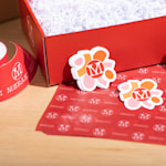 Two custom die-cut stickers shaped like a brand logo, displayed alongside branded roll labels, a mailer box and tissue paper.