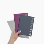 Two custom wraparound spiral notebooks in a person's hand, in maroon and navy. Both feature simple text on the right-hand edge of the cover.
