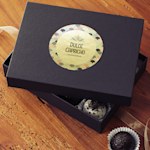 A black box sitting on a wooden table with the cover off revealing bon bons inside. The lid has a circular gold paper label applied to it.