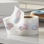 A roll of die-cut label stickers in the shape of two doughnuts in an offset stack placed on a countertop infront of two boughnut boxes, and a tray of doughnuts.