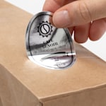 A bag of coffee on a gray background with a hand applying a silver paper oval label to the front.