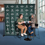 A man and a woman having coffee in front of a step & repeat banner with a logo and business name printed all over it.