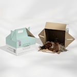 2 cajas loncheras personalizadas de una empresa de productos de belleza. 1 caja lonchera de lado con papel triturado y 2 botellas de productos adentro.