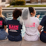 Un grupo pequeño de mujeres sentadas junto a una pileta que tienen puestas camisetas de viaje de despedida de soltera.