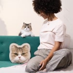 Una mujer está sentada en un sofá con su gato mientras mira un cojín con la cara de su peludo amigo impresa en él.