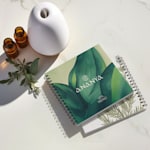 Catálogos con encuadernado de doble alambre de un spa y productos de bienestar con diseños botánicos de hojas, con un difusor y aceites esenciales a su alrededor.